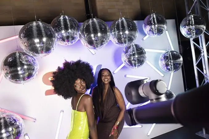 Two women posing under mirror balls in front of a robotic arm camera